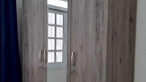 Ein großer Schrank mit zwei Türen und einem Spiegel, vor einer schlichten Wand mit Fenster und blauem Vorhang.
