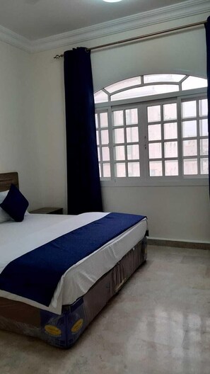 A bedroom with a bed, a window with curtains, and a view of buildings outside. - Today House Suites 2 (Salalah)