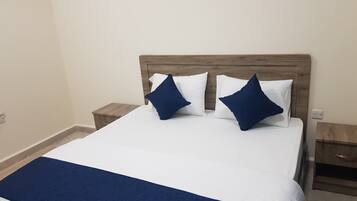 A neatly made bed with a wooden headboard, two white pillows, and two dark blue pillows. There are two wooden nightstands, one on each side of the bed.