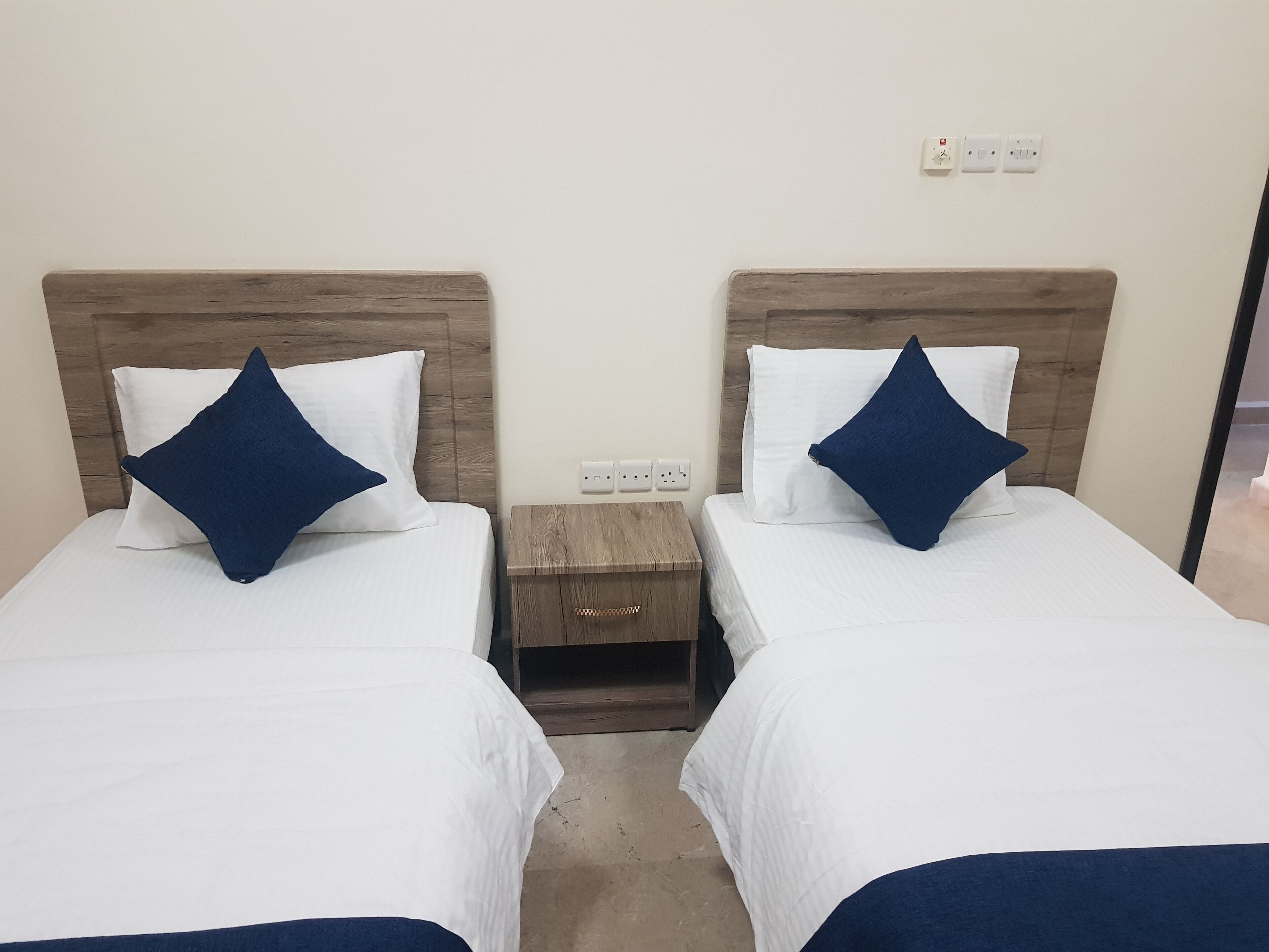Dos camas individuales con cabeceros de madera, ropa de cama blanca y almohadas azul oscuro. Hay una mesita de noche de madera entre las camas con un enchufe arriba.