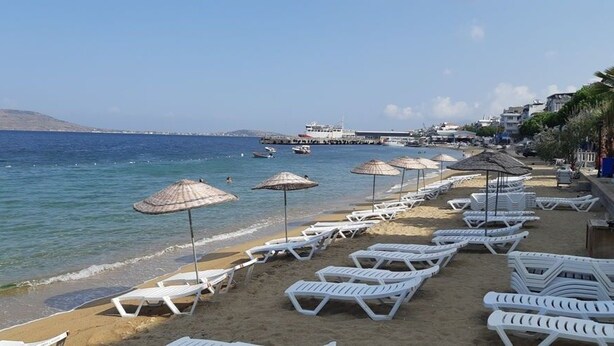A beach with white lounge chairs and thatched umbrellas, a pier in the background, and a clear blue sea. - Avşa Çakır Apart (Marmara)