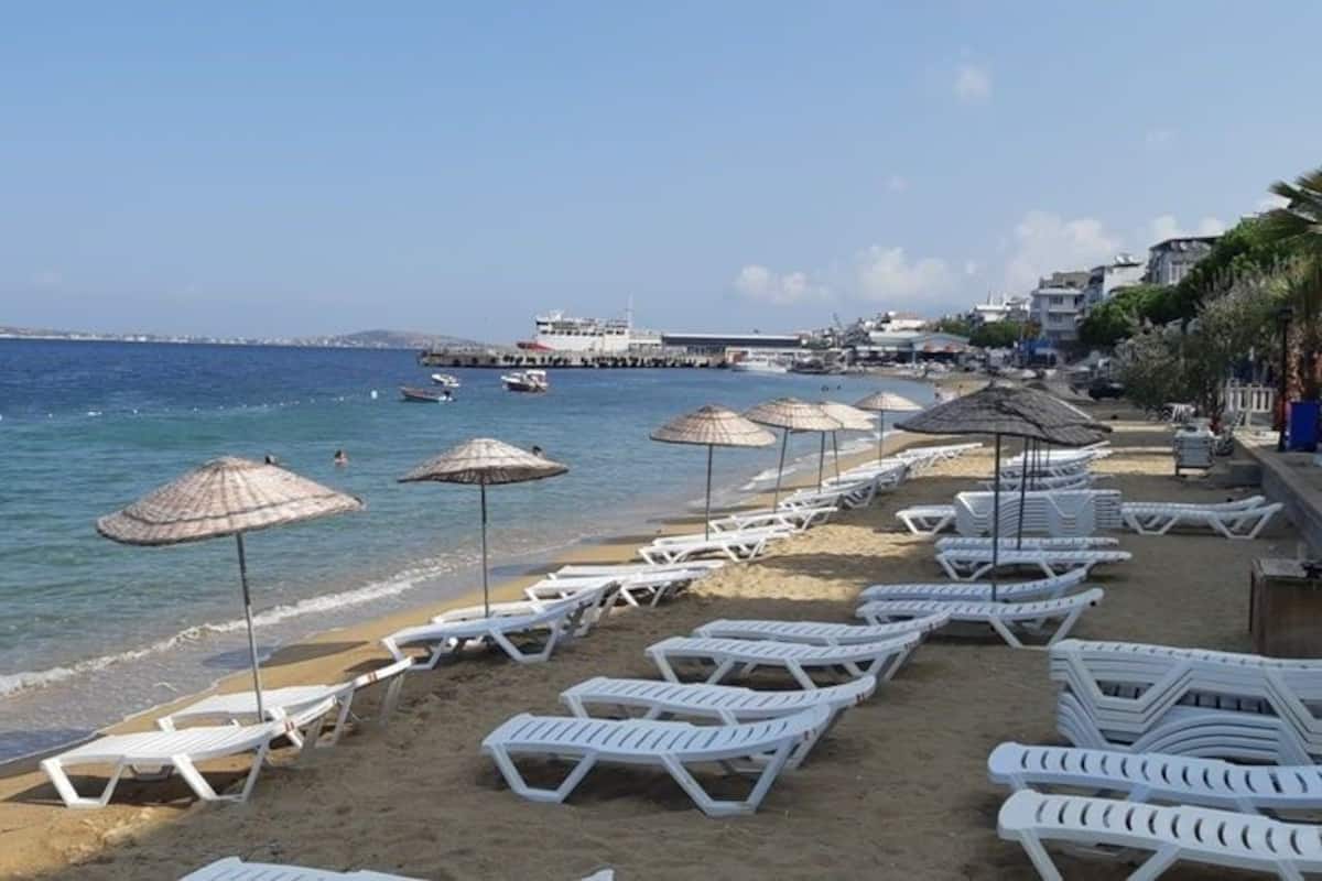 A beach with white lounge chairs and thatched umbrellas, a pier in the background, and a clear blue sea.
