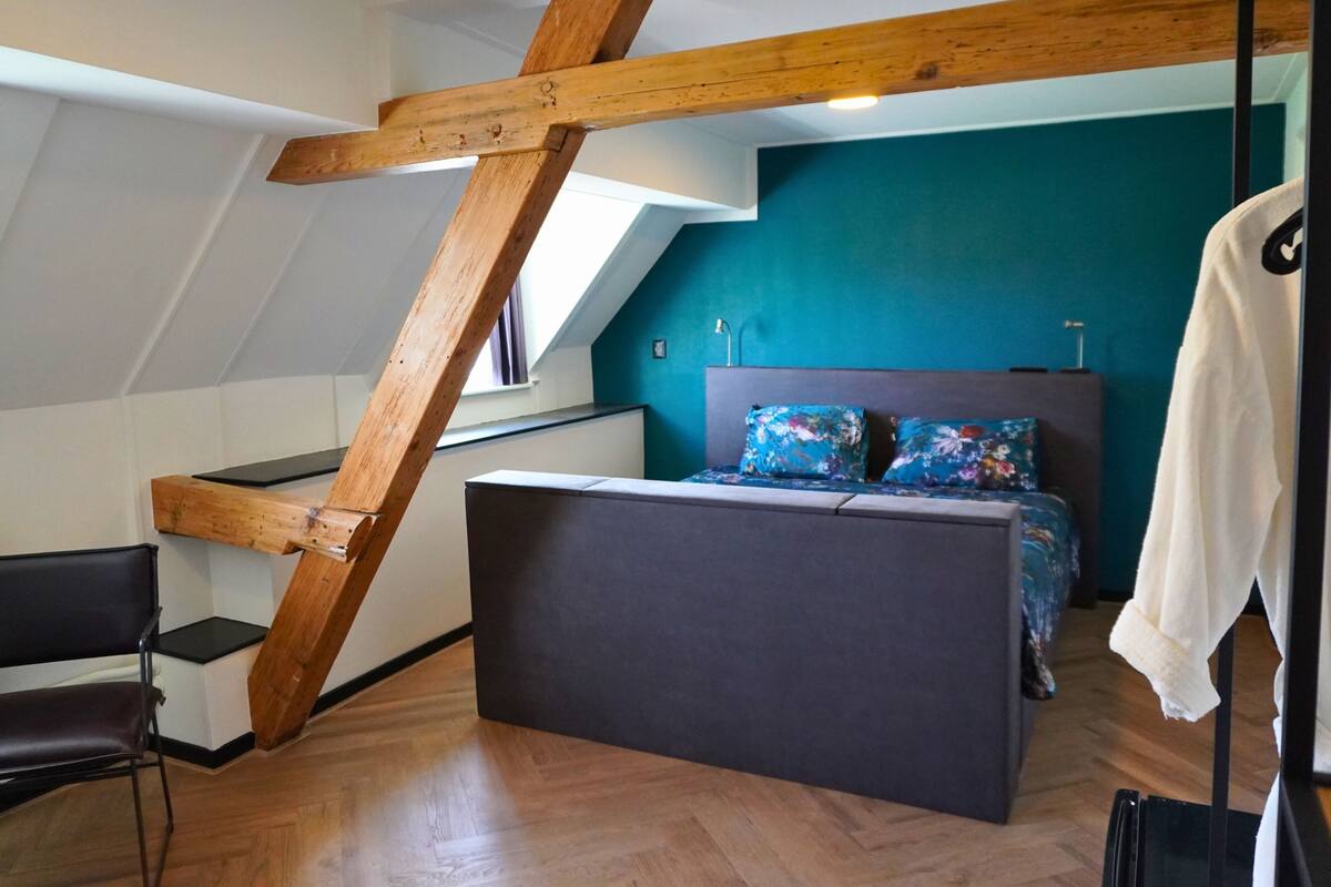 Une chambre avec un plafond en pente, un lit avec une literie à motifs floraux, une chaise et un porte-manteau.