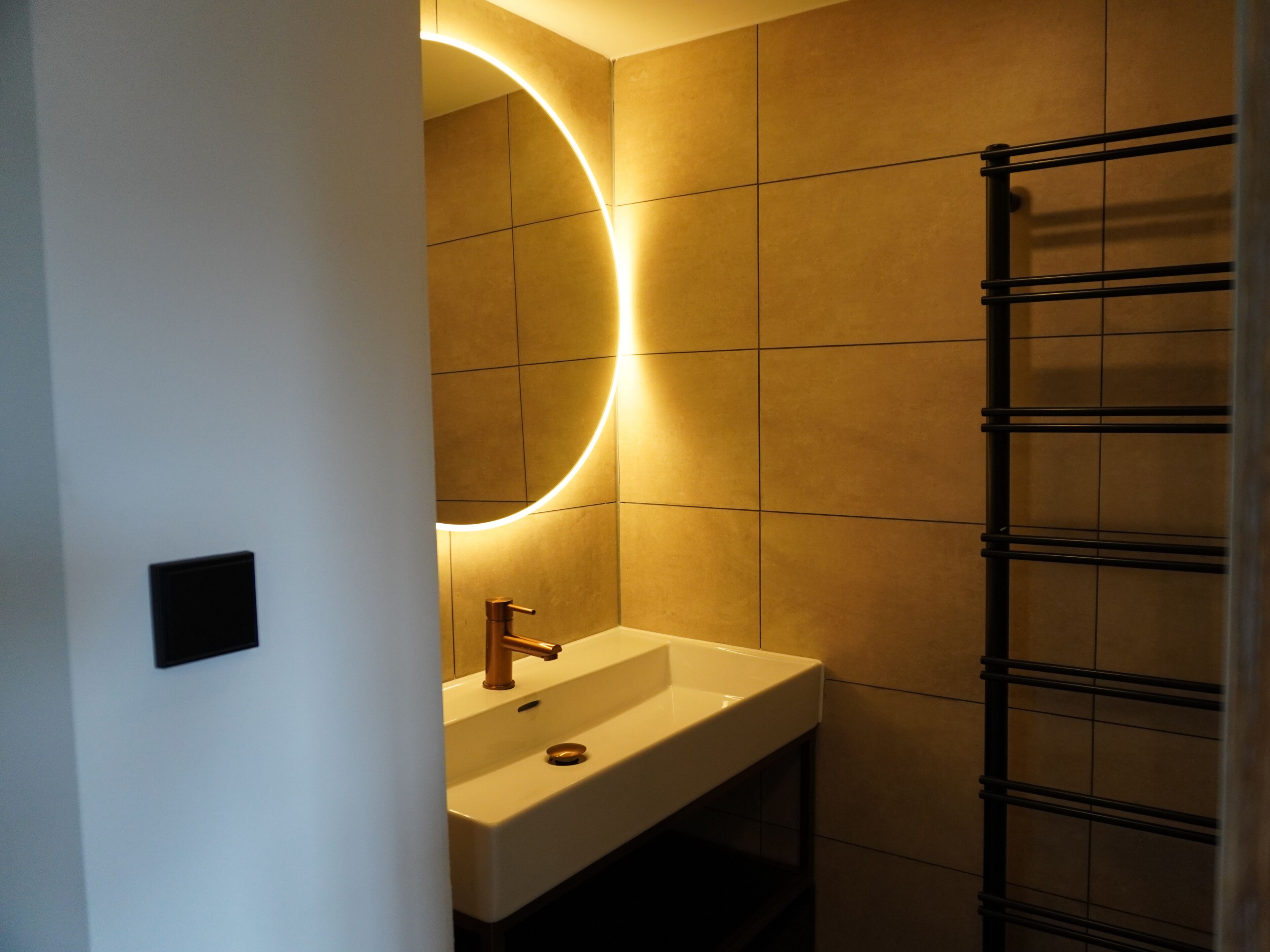 Een badkamer met een witte wastafel, een ronde spiegel met verlichting en een handdoekhouder.