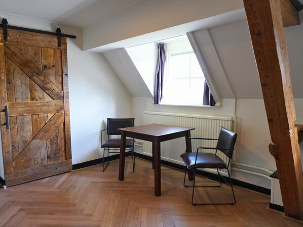 A room with a wooden table and chairs, a sliding barn door, a window with curtains, and a radiator. - B&B Catharinenburg (Melissant)