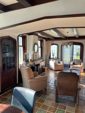A spacious living room with a fireplace, comfortable seating, and large windows offering natural light. - B&B Catharinenburg (Melissant)