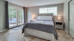 A bedroom with a bed, two bedside tables, a window, and a sliding glass door leading to an outdoor area. - Steps to Beach & Bay | A/C, Patio & 4 Park Spots (San Diego)