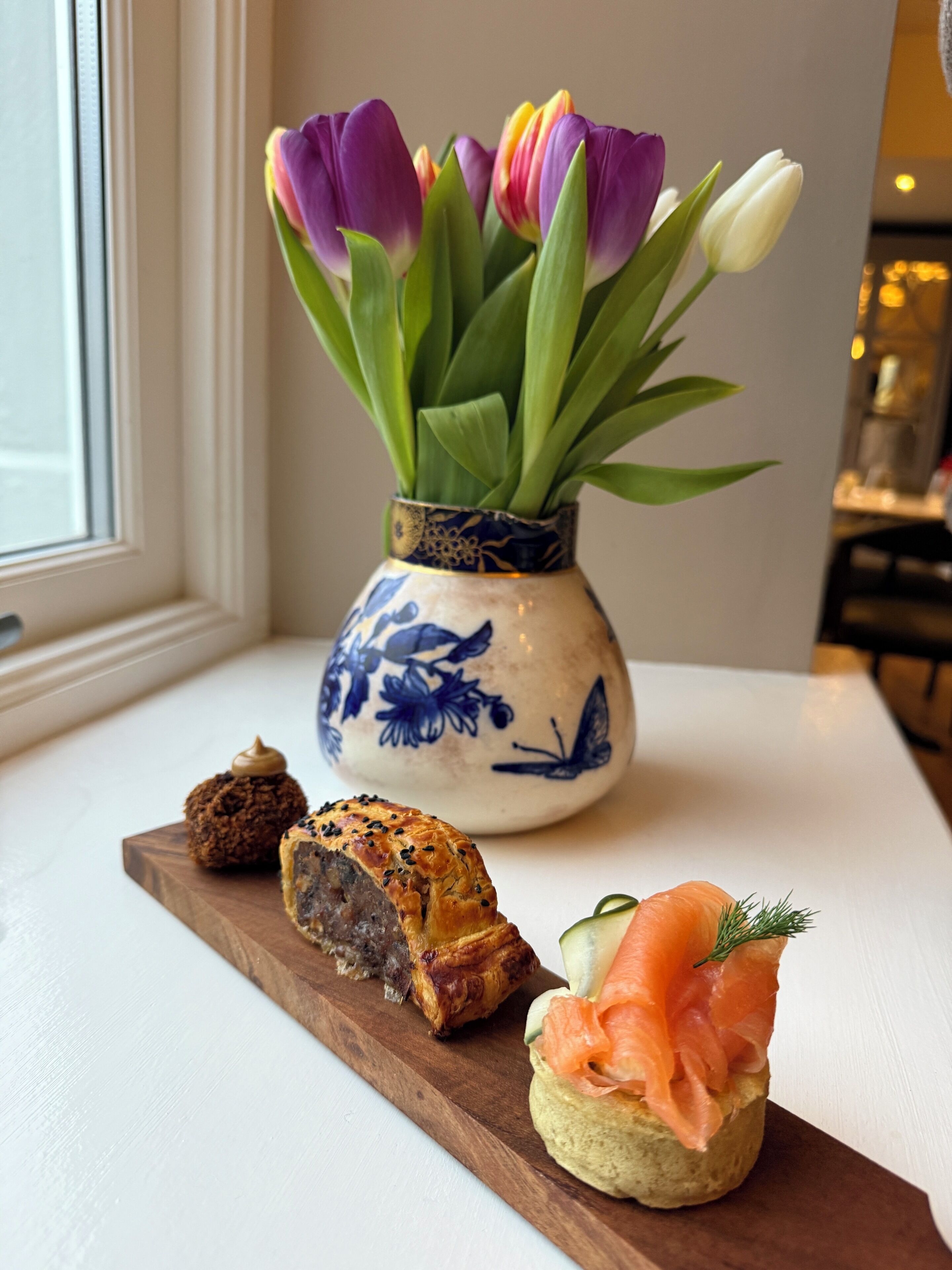 Een tafel met een bloemenvaas, een houten plank met gebak, en een bord gerookte zalm.