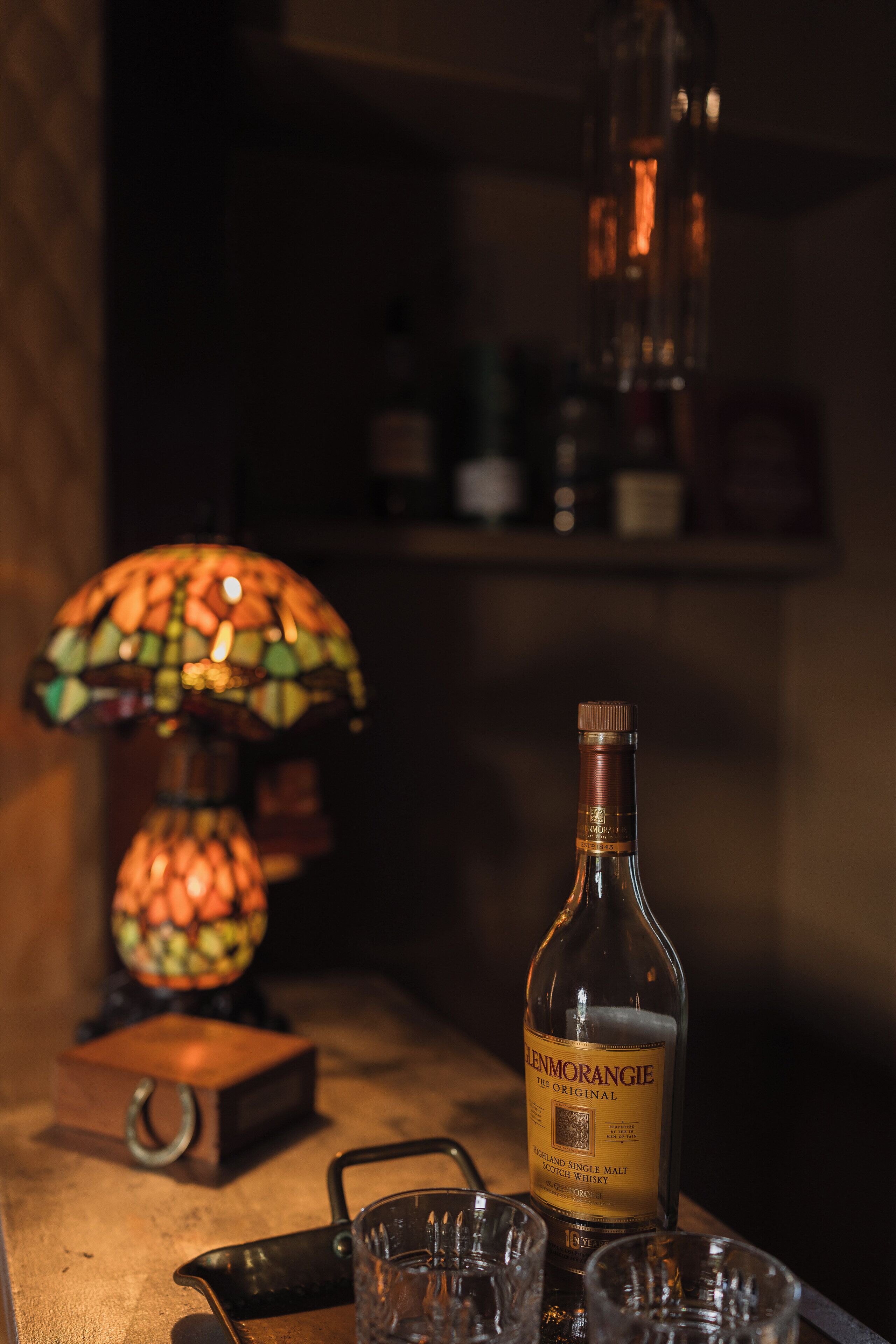 Una botella de whisky Glenmorangie sobre una mesa de madera, con una lámpara de vitral y una bandeja de metal con vasos.