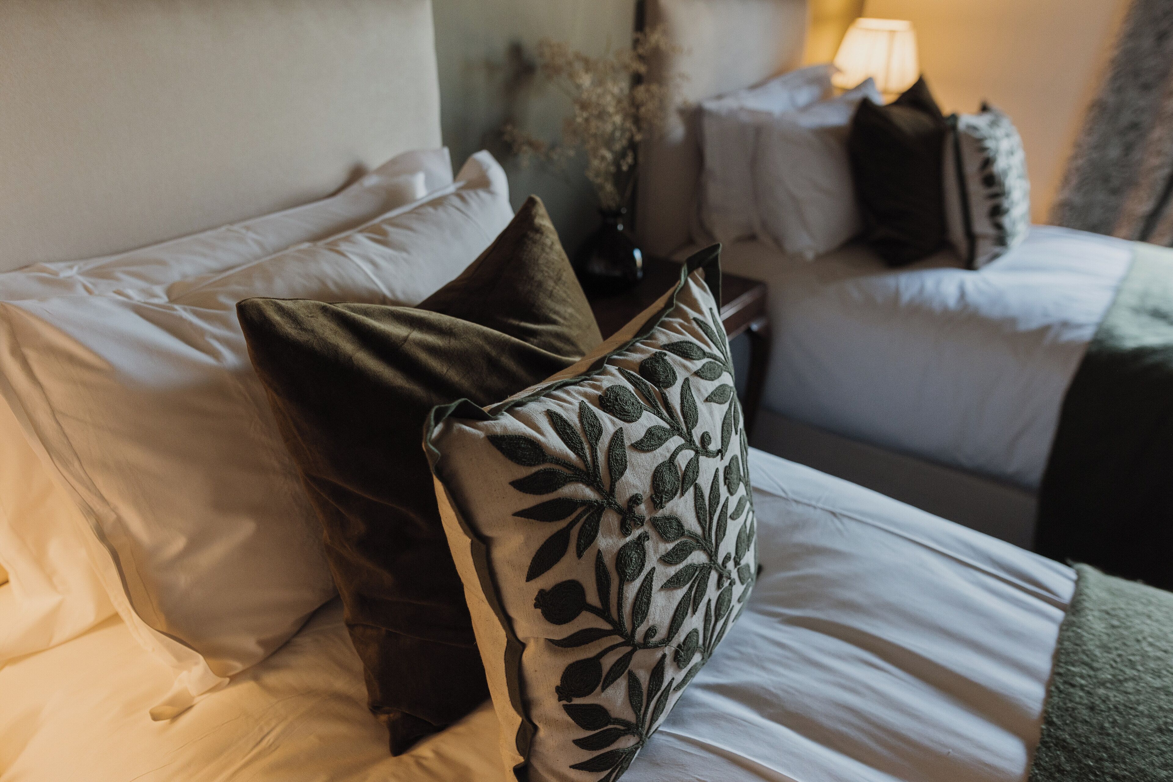 Una cama bien hecha con sábanas blancas y almohadas decorativas, una mesita de noche con lámpara, y un florero con flores secas.