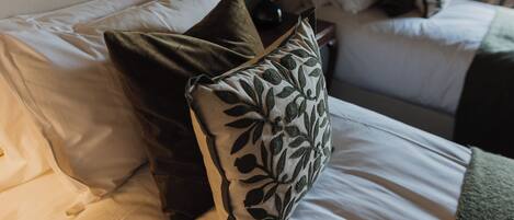 Una cama bien hecha con sábanas blancas y almohadas decorativas, una mesita de noche con lámpara, y un florero con flores secas.