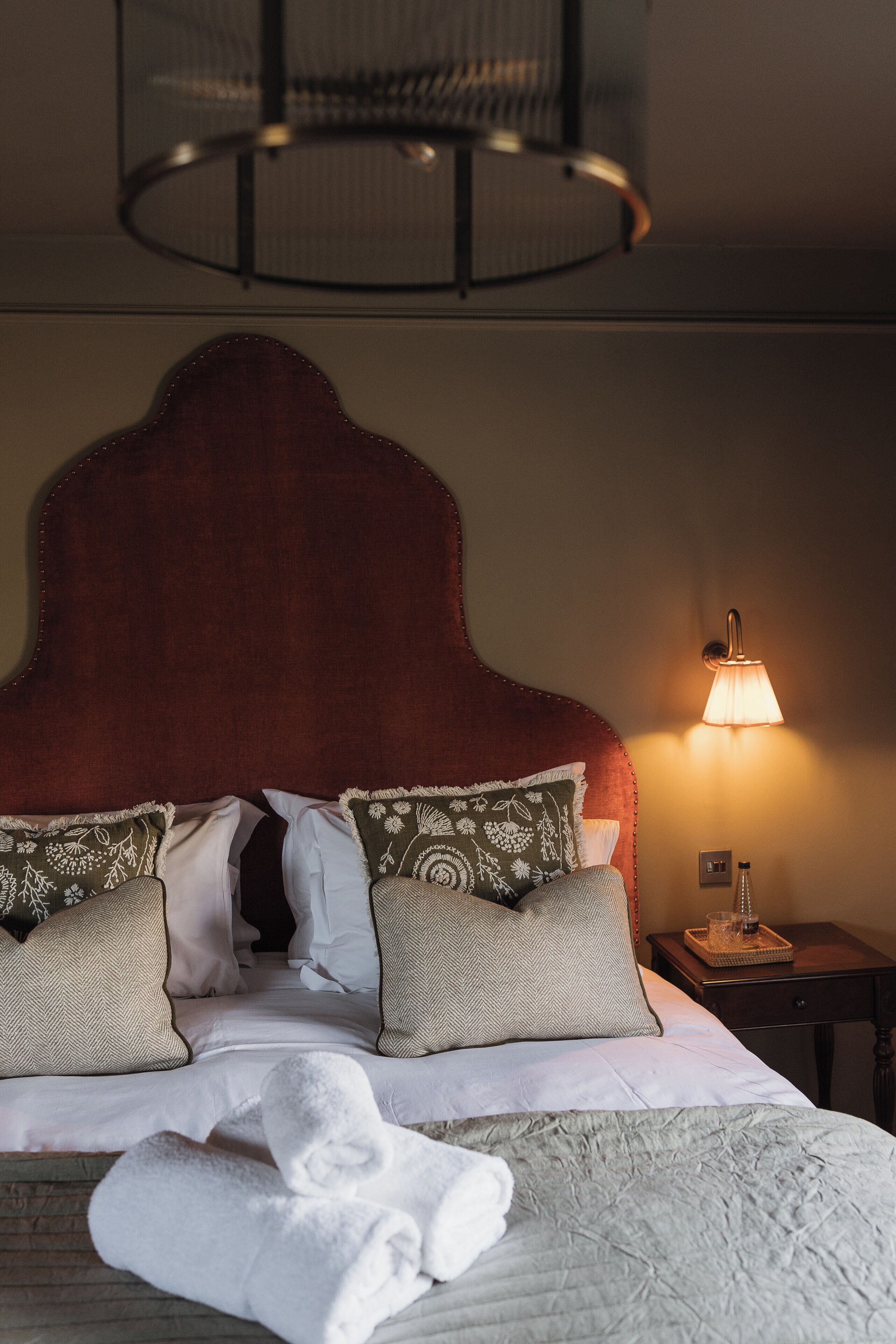 Un lit bien fait, avec une tête de lit rouge, du linge de lit blanc et des coussins décoratifs. On aperçoit une lampe de chevet et une petite table sur laquelle sont posés un verre et un livre.