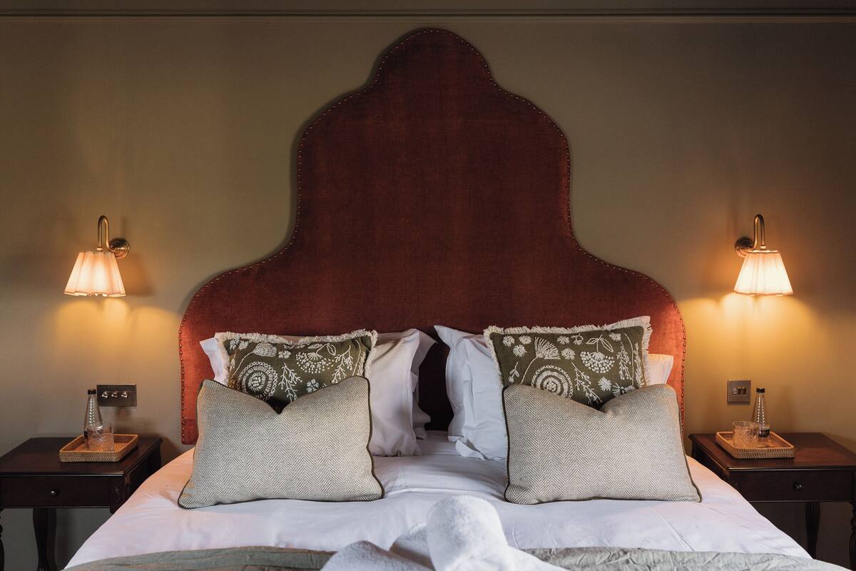 Una cama bien hecha con un cabecero tapizado en rojo, dos mesitas de noche con lámparas y almohadas decorativas.