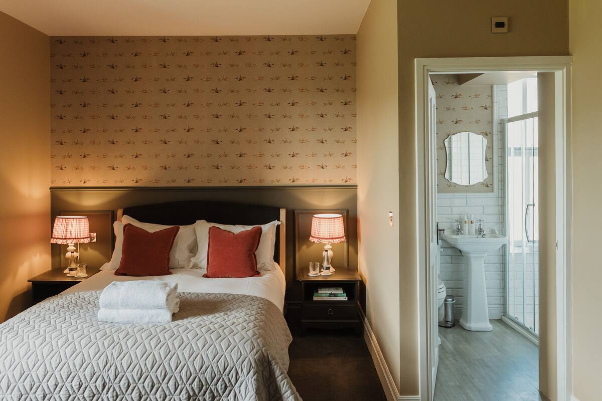 Una habitación de hotel con una cama, dos mesitas de noche con lámparas, una mesita auxiliar, una almohada roja, una almohada blanca, una toalla doblada, una pequeña mesa con libros y un baño visible a través de una puerta abierta.