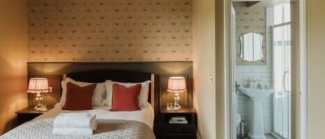 Una habitación de hotel con una cama, dos mesitas de noche con lámparas, una mesita auxiliar, una almohada roja, una almohada blanca, una toalla doblada, una pequeña mesa con libros y un baño visible a través de una puerta abierta.