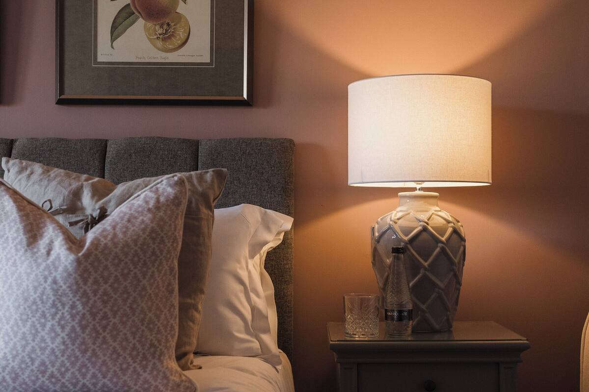 Una cama bien hecha con almohadones estampados, una mesita de noche con lámpara e impresión botánica enmarcada en la pared.