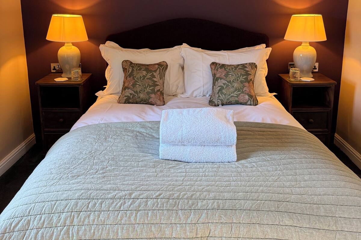 Una cama bien hecha con una colcha acolchada, dos almohadas y una toalla doblada. Hay dos lámparas de noche sobre mesitas de madera, y un cuadro enmarcado cuelga sobre la cama.