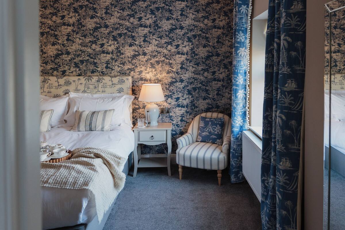 Un dormitorio con cama, mesita de noche, lámpara, silla y ventana con cortinas.