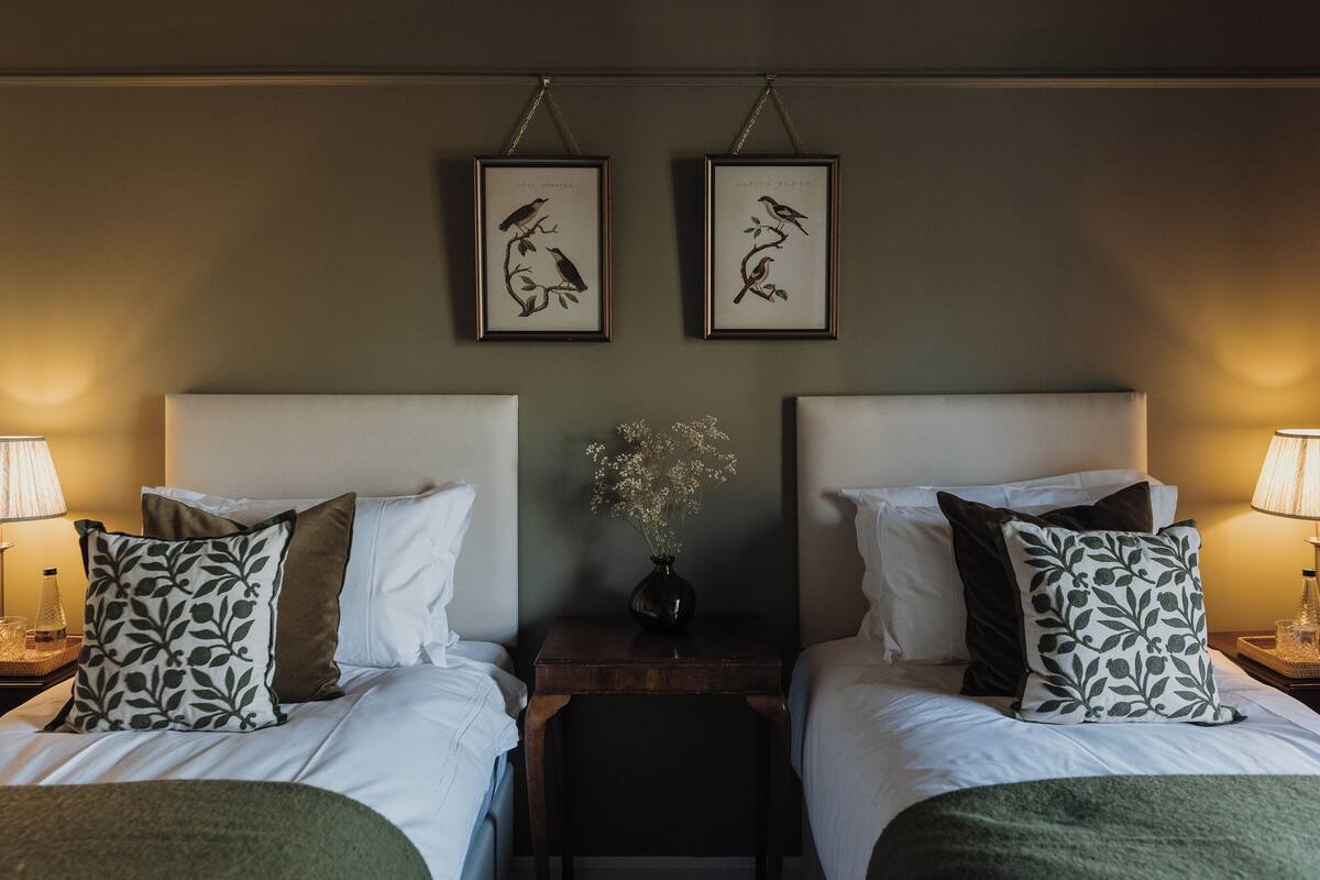 Habitación de hotel con dos camas, ropa de cama blanca, almohadas con estampado oscuro, una mesita de noche con un jarrón de flores y cuadros en la pared.