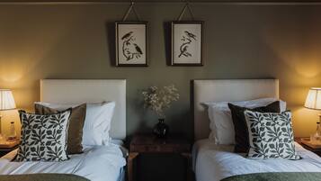 Une chambre d’hôtel avec deux lits, du linge de lit blanc, des coussins à motifs foncés, une table de chevet avec un vase de fleurs et des œuvres d’art encadrées accrochées au mur.