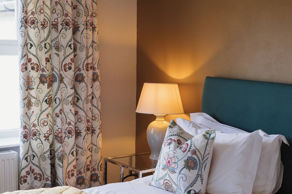 Una habitación de hotel con cama, mesita de noche, lámpara y ventana con cortinas.