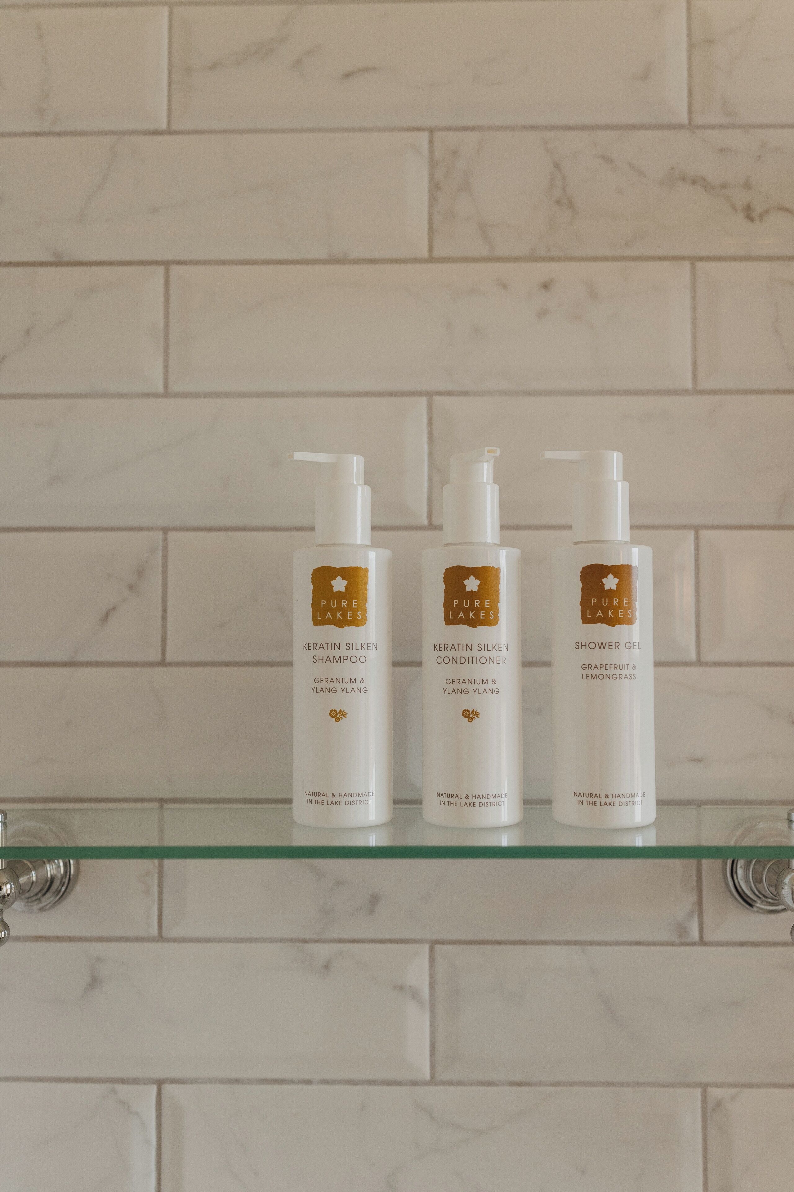 Tres botellas de productos Pure Lakes sobre un estante de vidrio frente a una pared de azulejos.