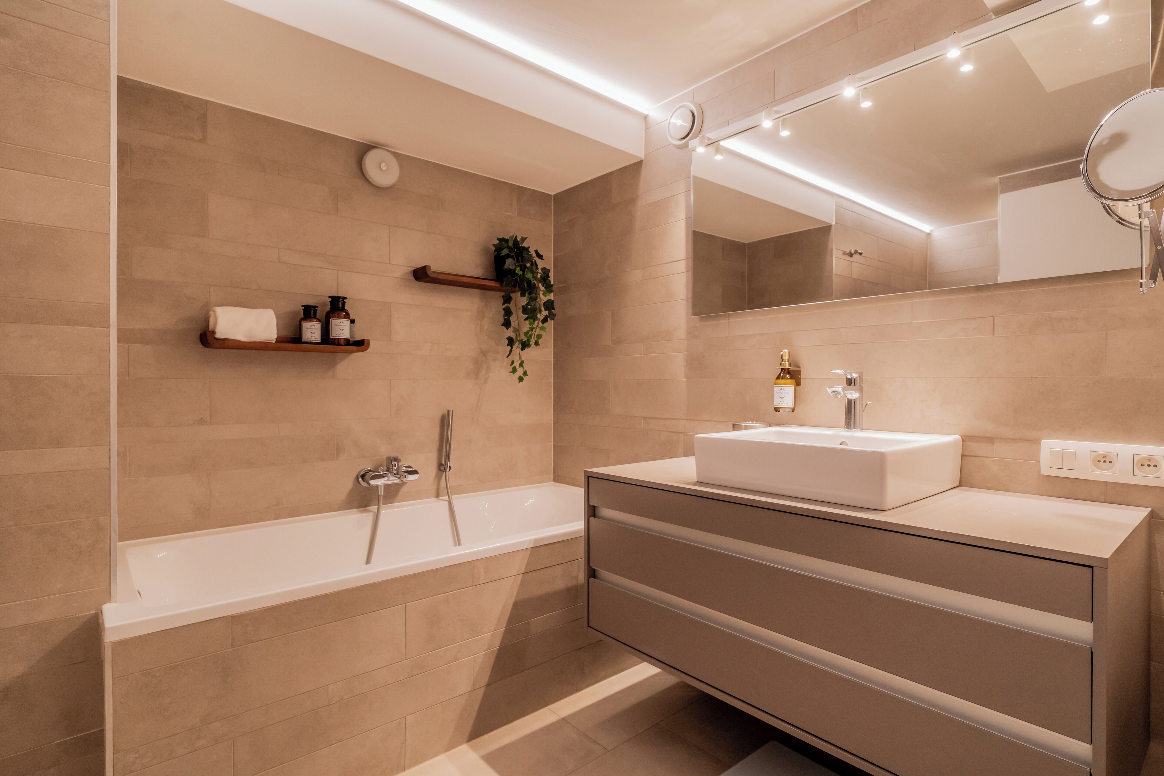 A modern bathroom with a bathtub, a vanity with a sink, and a mirror. There is a shelf with toiletries and a plant.