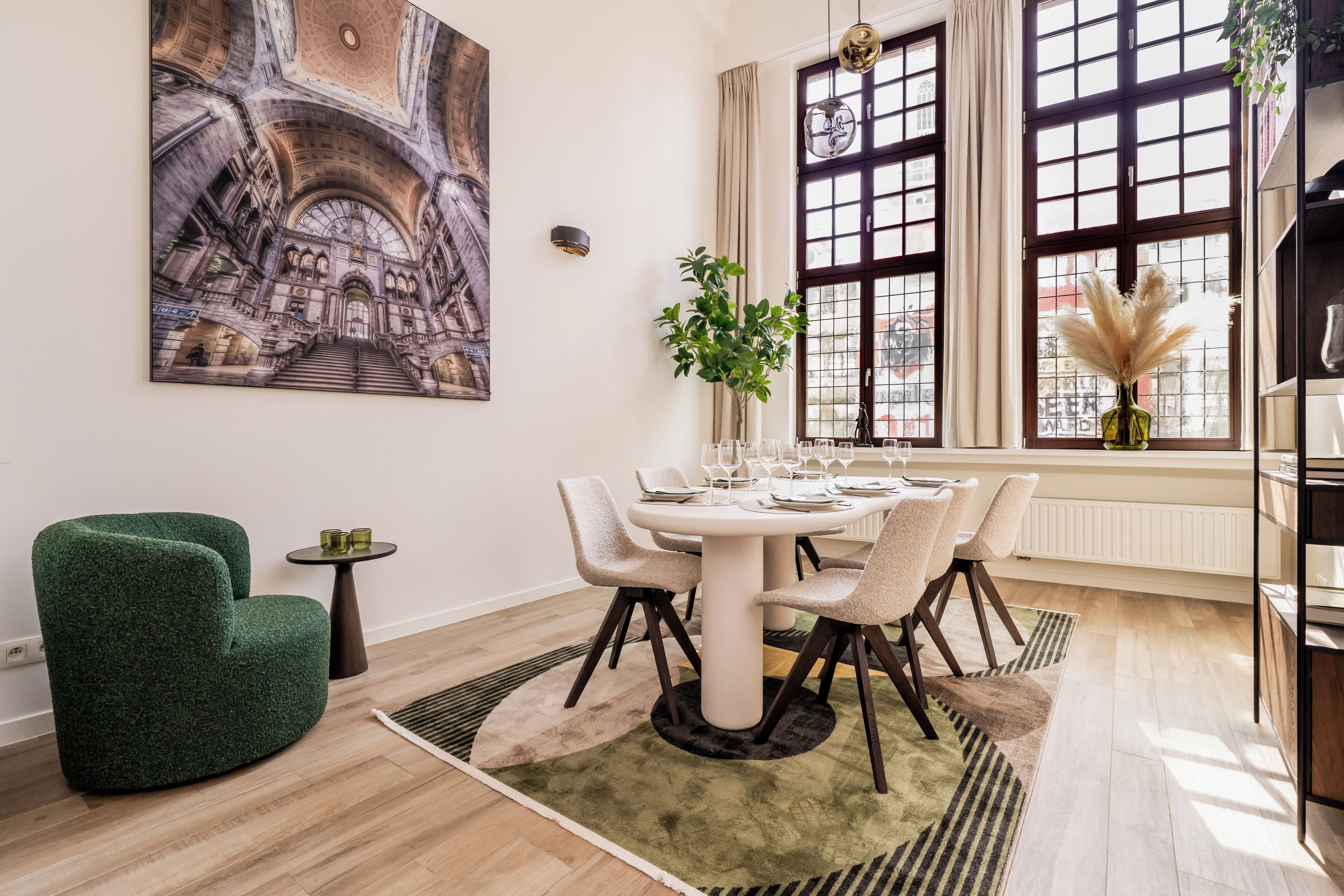 A dining area with a round table, chairs, a green armchair, a small side table, and a large painting on the wall.