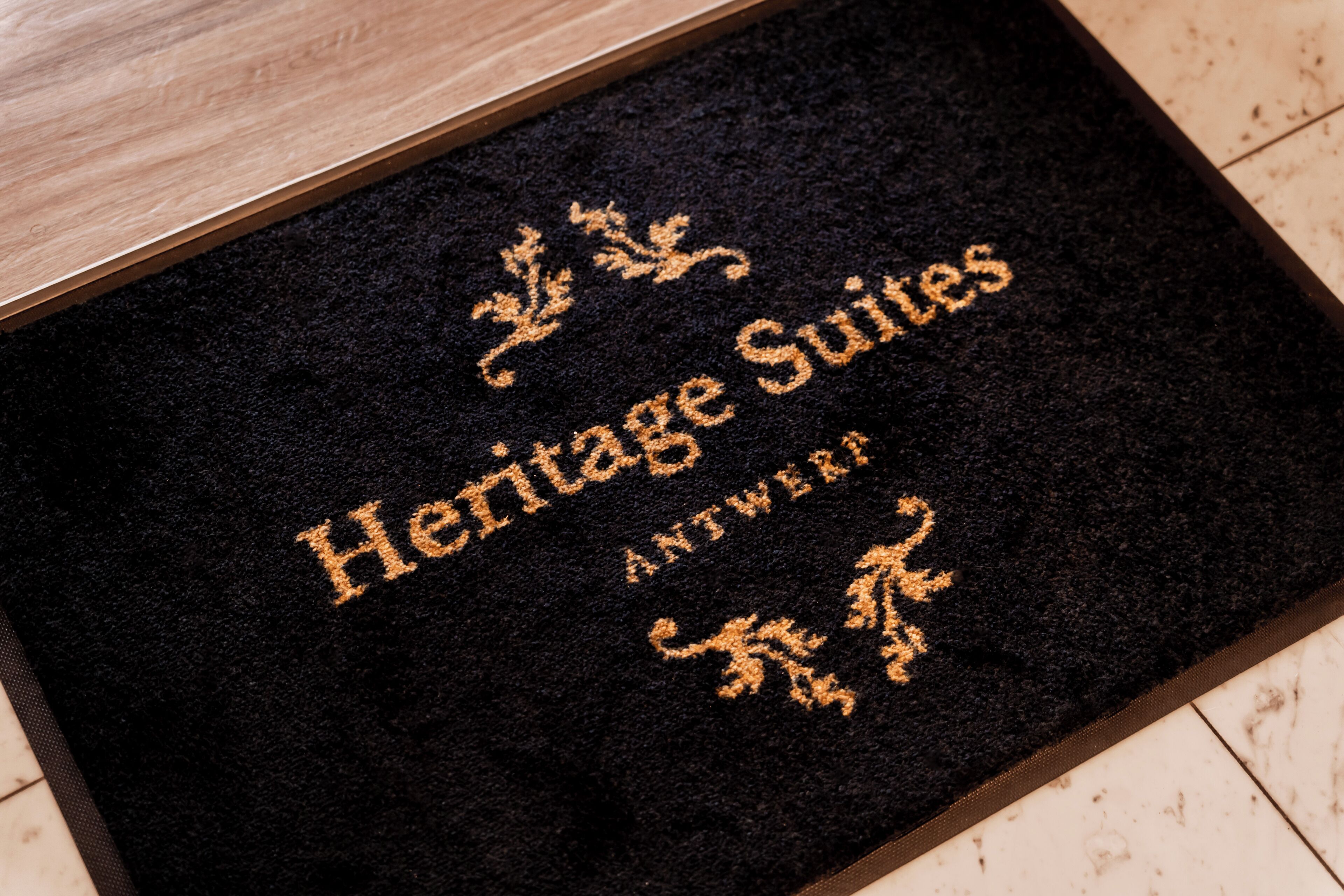 Een zwarte deurmat met in goud reliëf de tekst 'Heritage Suites' en 'Antwerpen'.