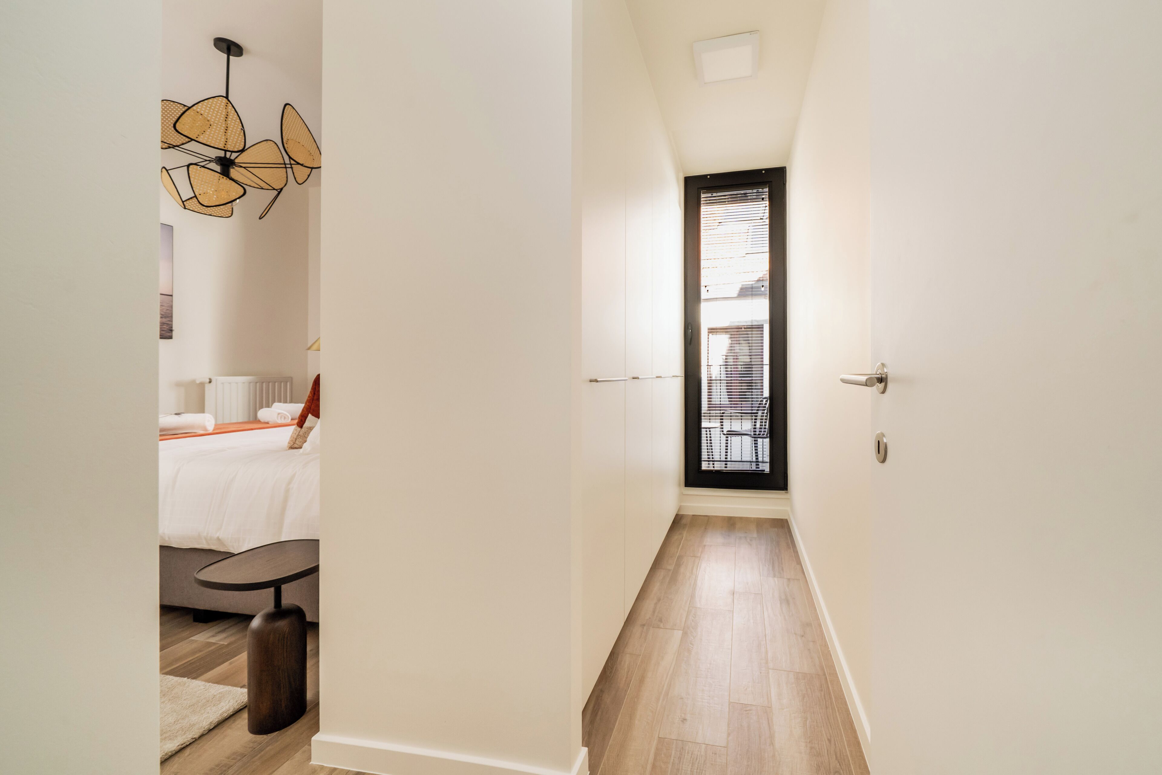 A narrow hallway with a bed, a table, and a chair in a room.