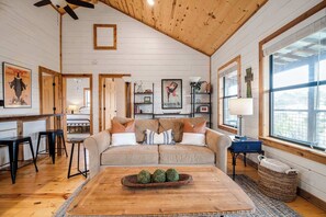 A cozy living room with a wooden ceiling, a sofa with cushions, a coffee table with decorative balls, and a view through large windows.