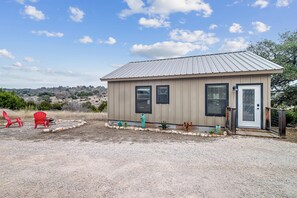 A small building with a metal roof, a front door, and two windows. There are red chairs and a small garden area with plants and rocks. - Modern Ranch Home with Hill Country Views (Gillespie County)
