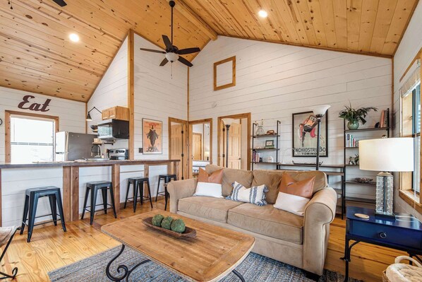 A cozy interior with a kitchen, dining area, and living room, featuring wooden walls, a ceiling fan, and a sofa.