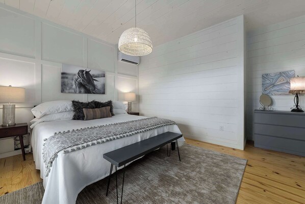 A bedroom with a bed, bedside lamps, a bench, a rug, a wall-mounted air conditioner, and a ceiling light fixture.