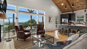 A spacious living area with a large window, a kitchen with a bar, and a balcony with outdoor furniture. - Modern Ranch Retreat with Hill Country Views (Gillespie County)