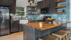 A modern kitchen with dark wood cabinets, a large island with stools, a stainless steel refrigerator, and a blue tiled backsplash. - Modern Ranch Retreat with Hill Country Views (Gillespie County)
