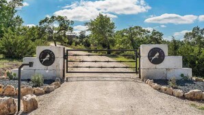 A gated entrance with a sign, a gravel path, and surrounding vegetation. - Modern Ranch Retreat with Hill Country Views (Gillespie County)