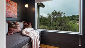 A room with a dark wooden wall, a gray sofa with patterned cushions, a large window with a view of trees, and a hanging light fixture. - Treehouse Retreat with Hill Country Views (Gillespie County)