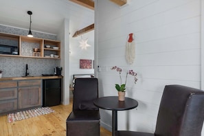 A modern kitchen with wooden shelves, a black refrigerator, and a microwave. There is a black leather sofa and a small round table with a potted orchid.
