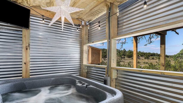 A hot tub in a rustic cabin with a star decoration, wooden beams, and a view of the desert landscape. - Treehouse Retreat with Hill Country Views (Gillespie County)