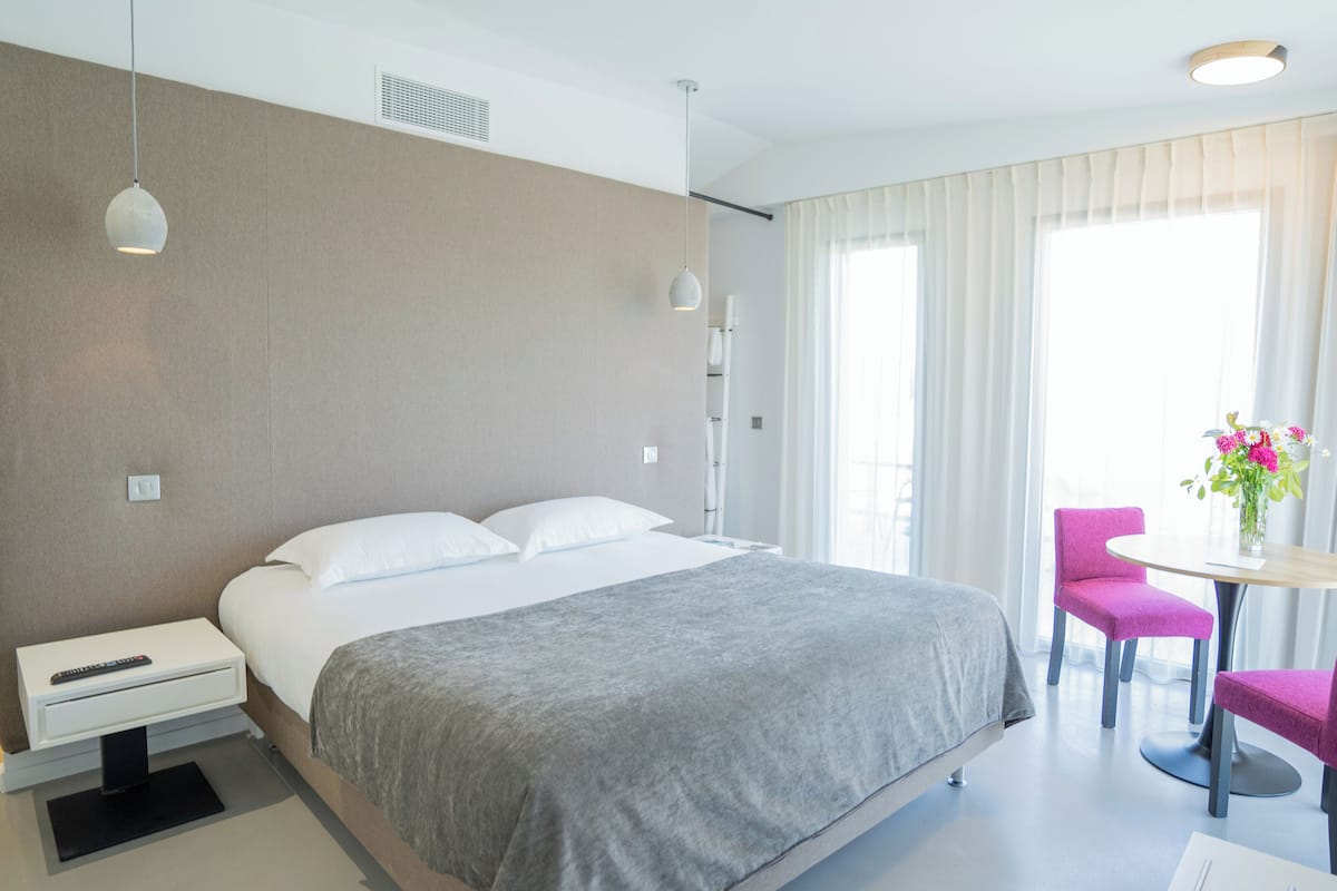 Ein modernes Hotelzimmer mit einem großen Bett, einem kleinen Tisch mit zwei Stühlen, einer Fernbedienung auf dem Nachttisch und einem Fenster mit durchsichtigen Vorhängen.