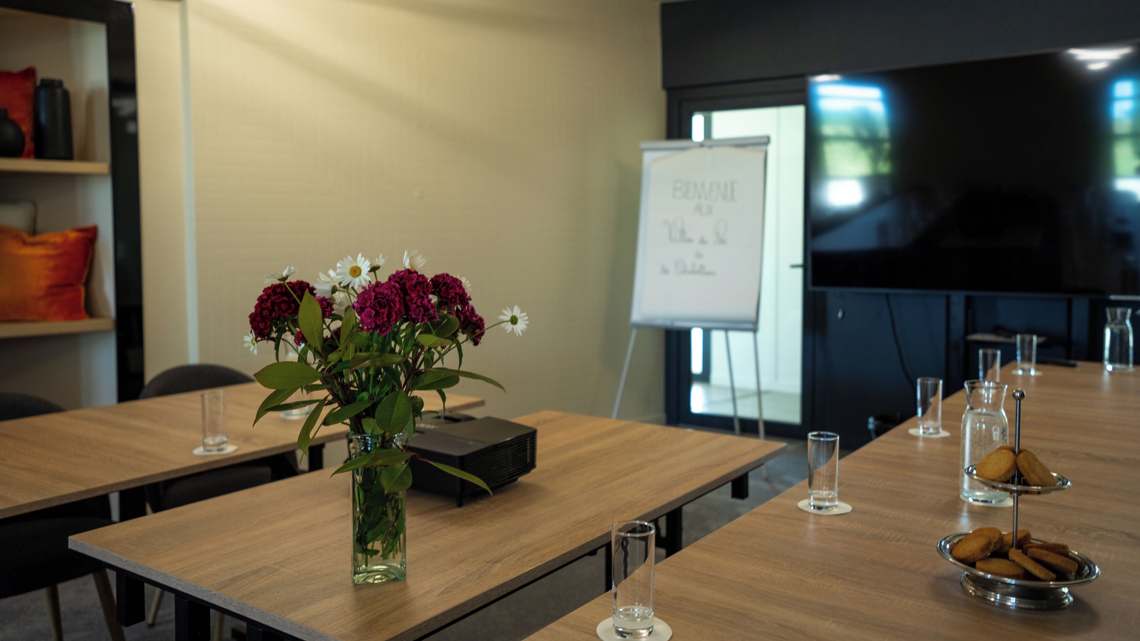 Un salón de conferencias con una larga mesa, un proyector, una pizarra y un jarrón con flores.