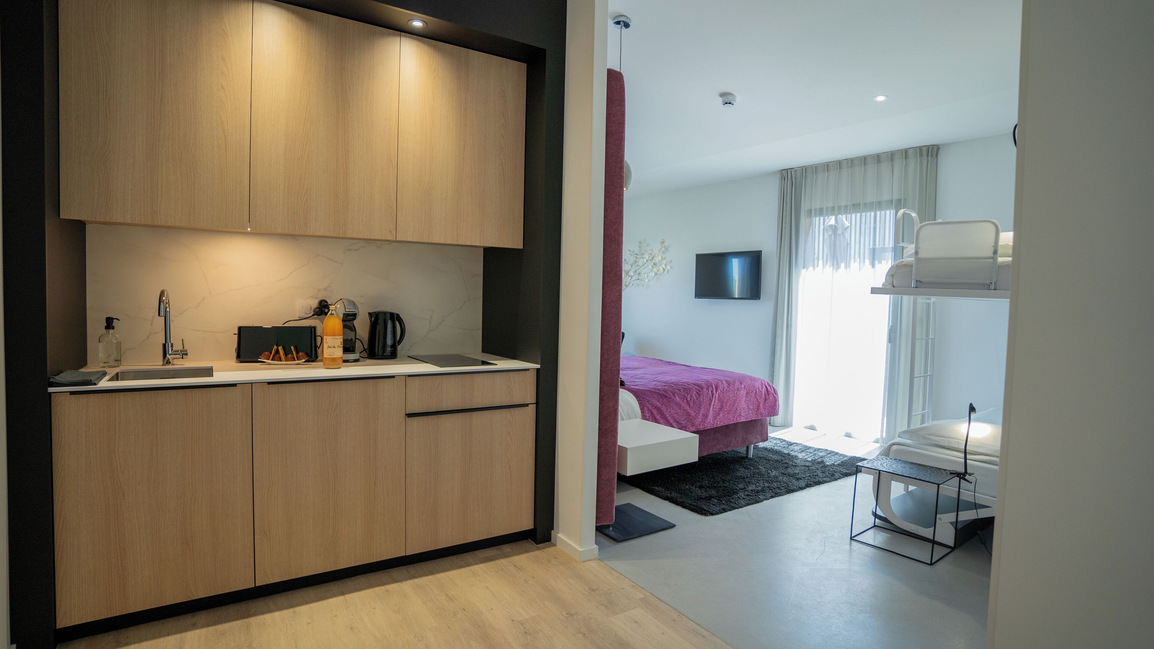 A modern hotel room with a kitchenette, a bed with a red bedspread, a TV mounted on the wall, and a desk with a chair.