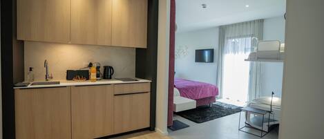A modern hotel room with a kitchenette, a bed with a red bedspread, a TV mounted on the wall, and a desk with a chair.