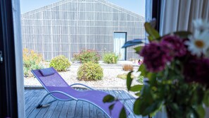 A modern building with a vertical striped facade, a wooden deck with a purple lounge chair, and various potted plants and shrubs in the foreground.