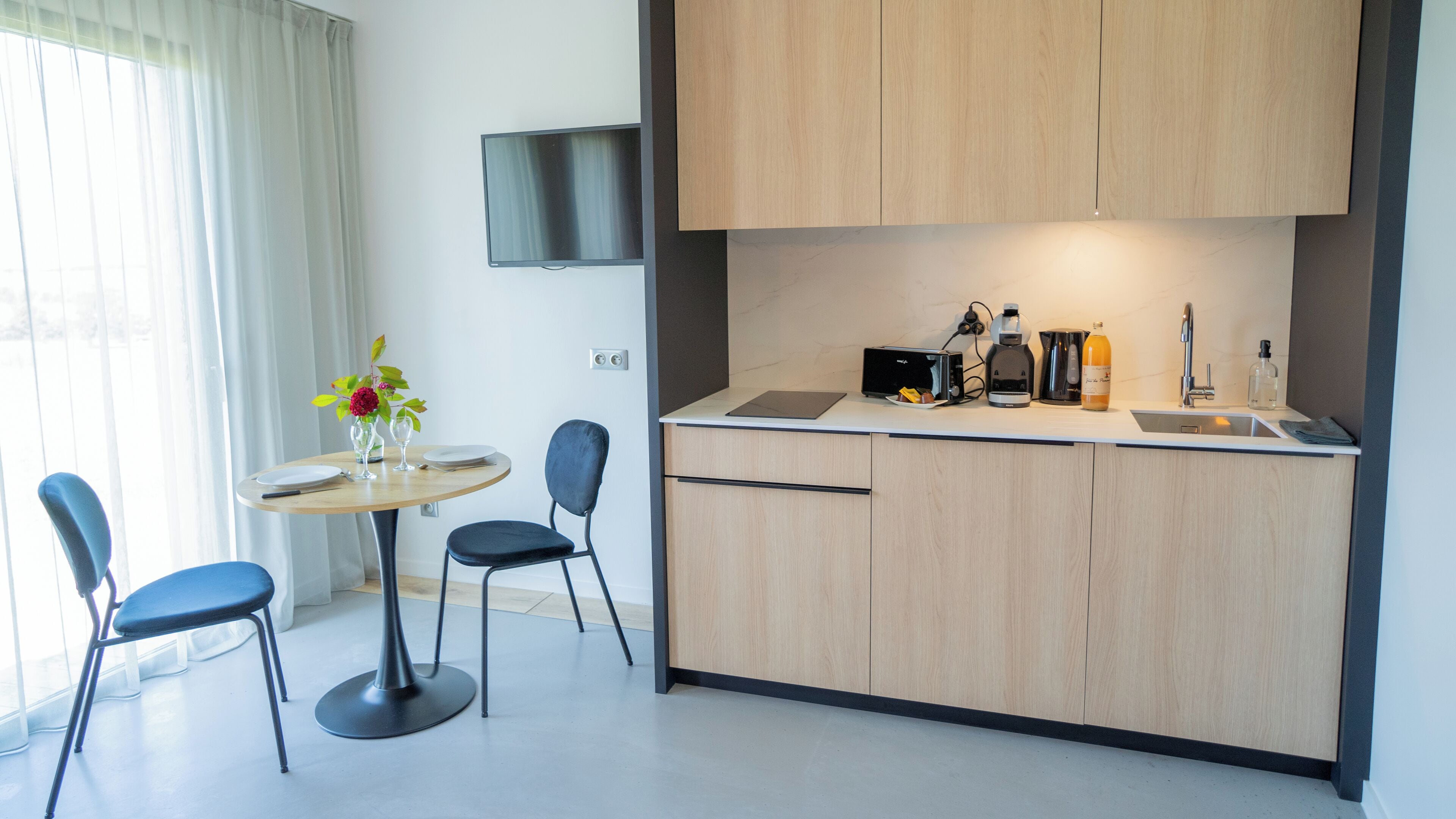 A modern kitchen with a dining area, including a table with chairs, a TV mounted on the wall, and various kitchen appliances.