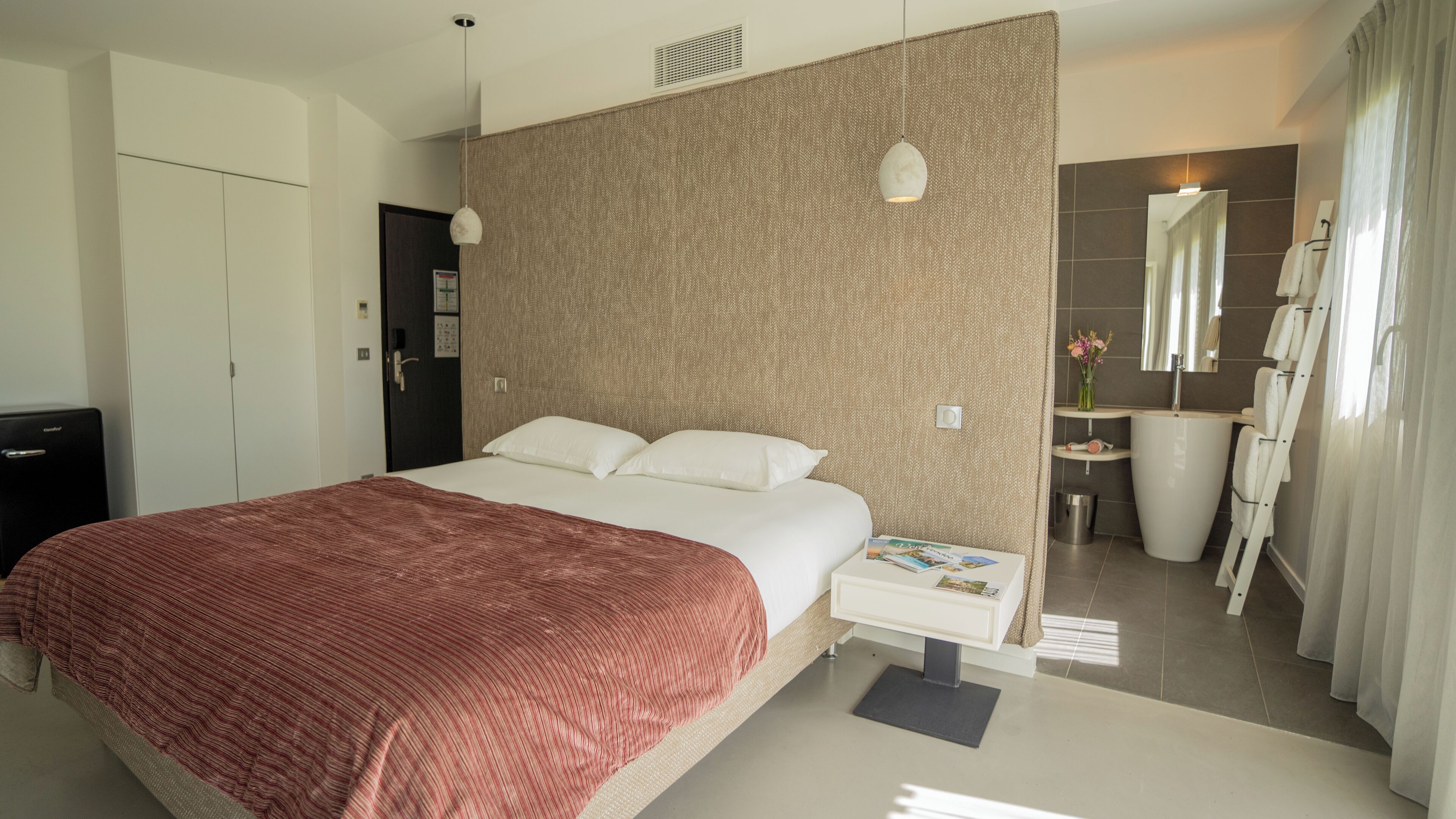 Habitación de hotel con una cama grande, una mesita de noche con una revista, un armario, un baño con bañera y zona de ducha.