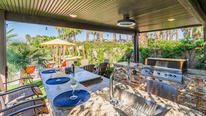 Un área de cocina exterior con una parrilla, taburetes de bar y una mesa de comedor.
