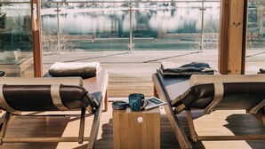 A modern outdoor seating area with two lounge chairs, a small wooden table, and a view of a snowy landscape through large windows.