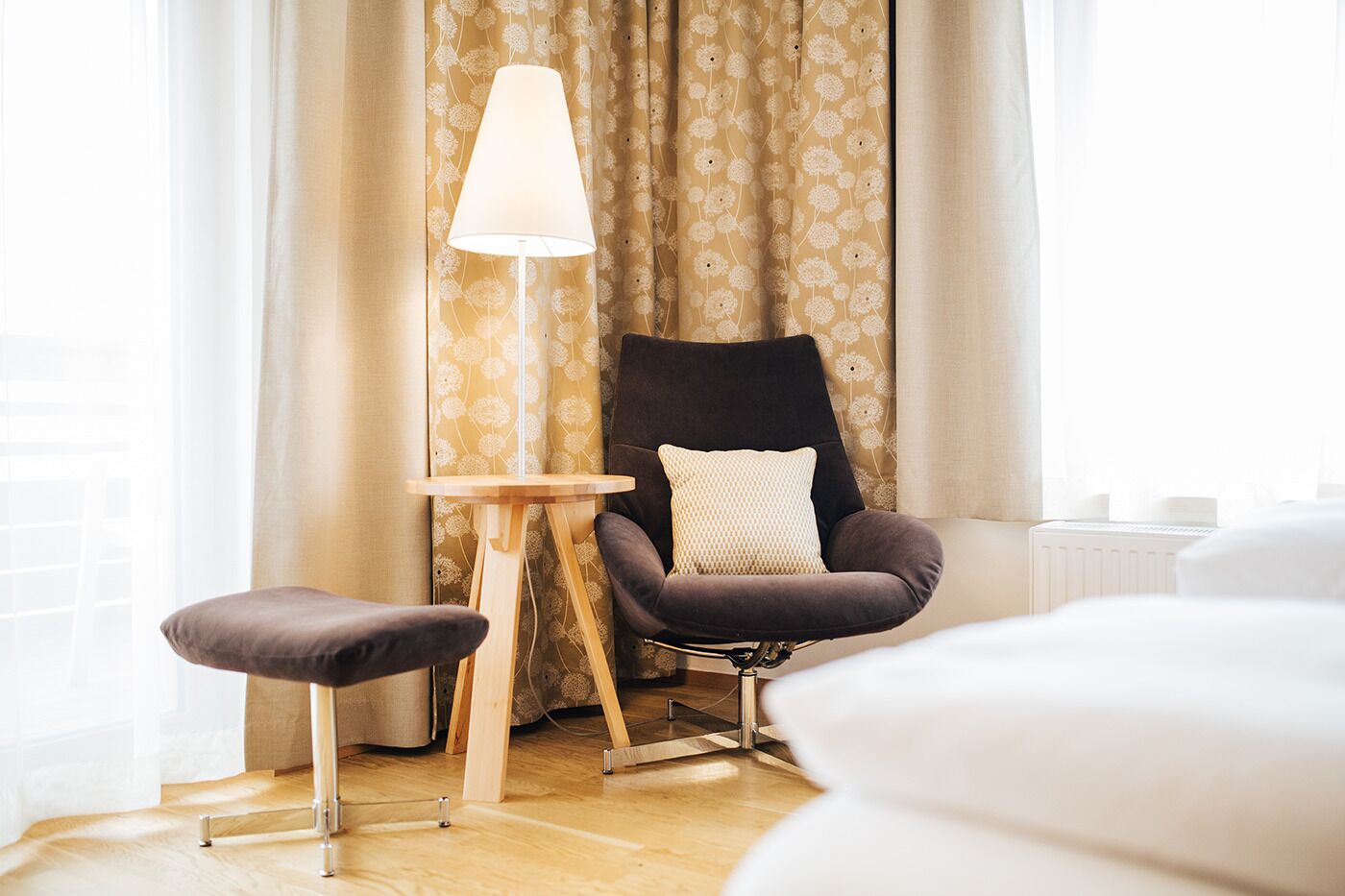 Ein modernes Hotelzimmer mit Holzboden, einem schwarzen Sessel mit Kissen, einem kleinen Holztisch und einer Stehlampe.